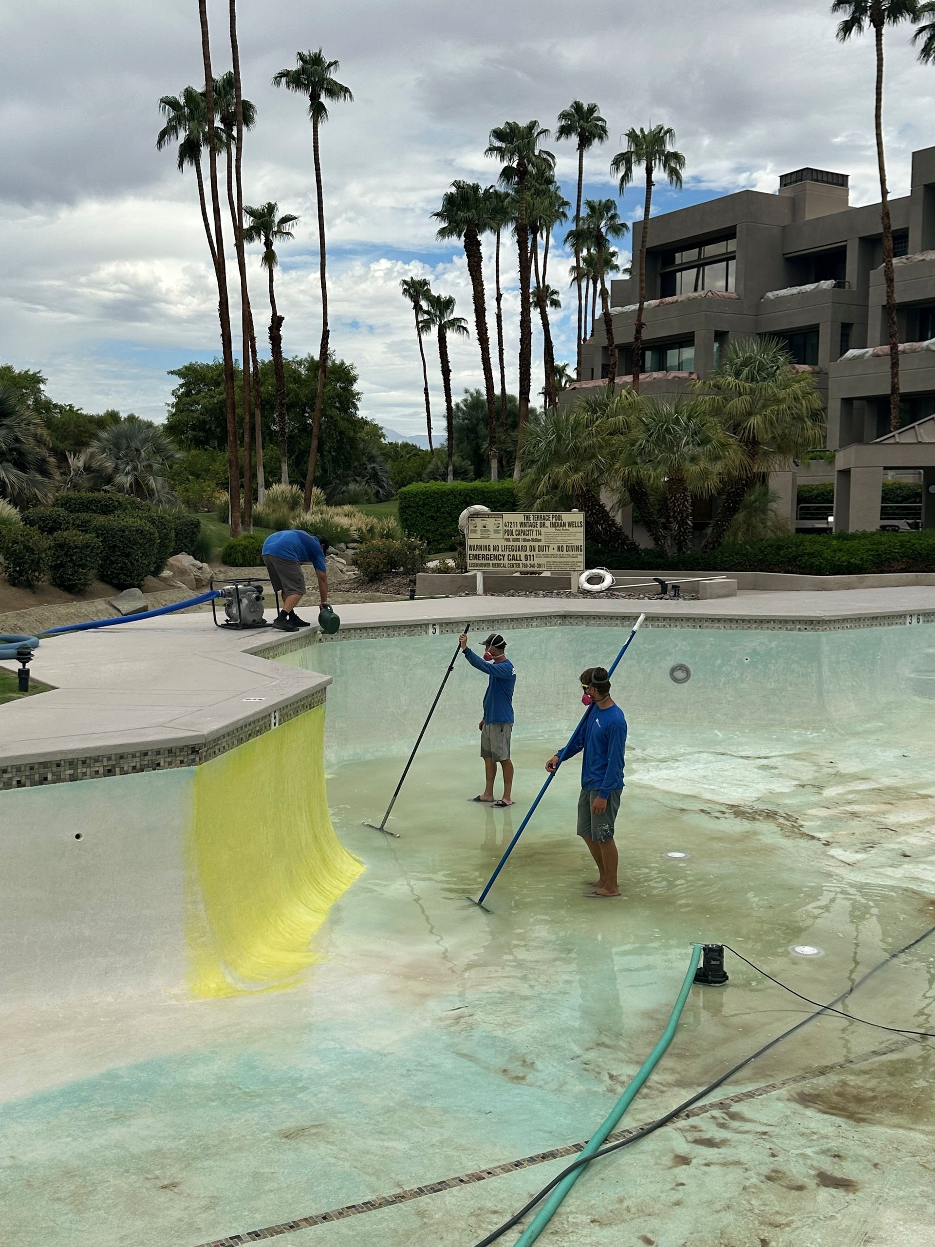 Children playing with hoses near a shallow resort pool, with palm trees and a building in the background