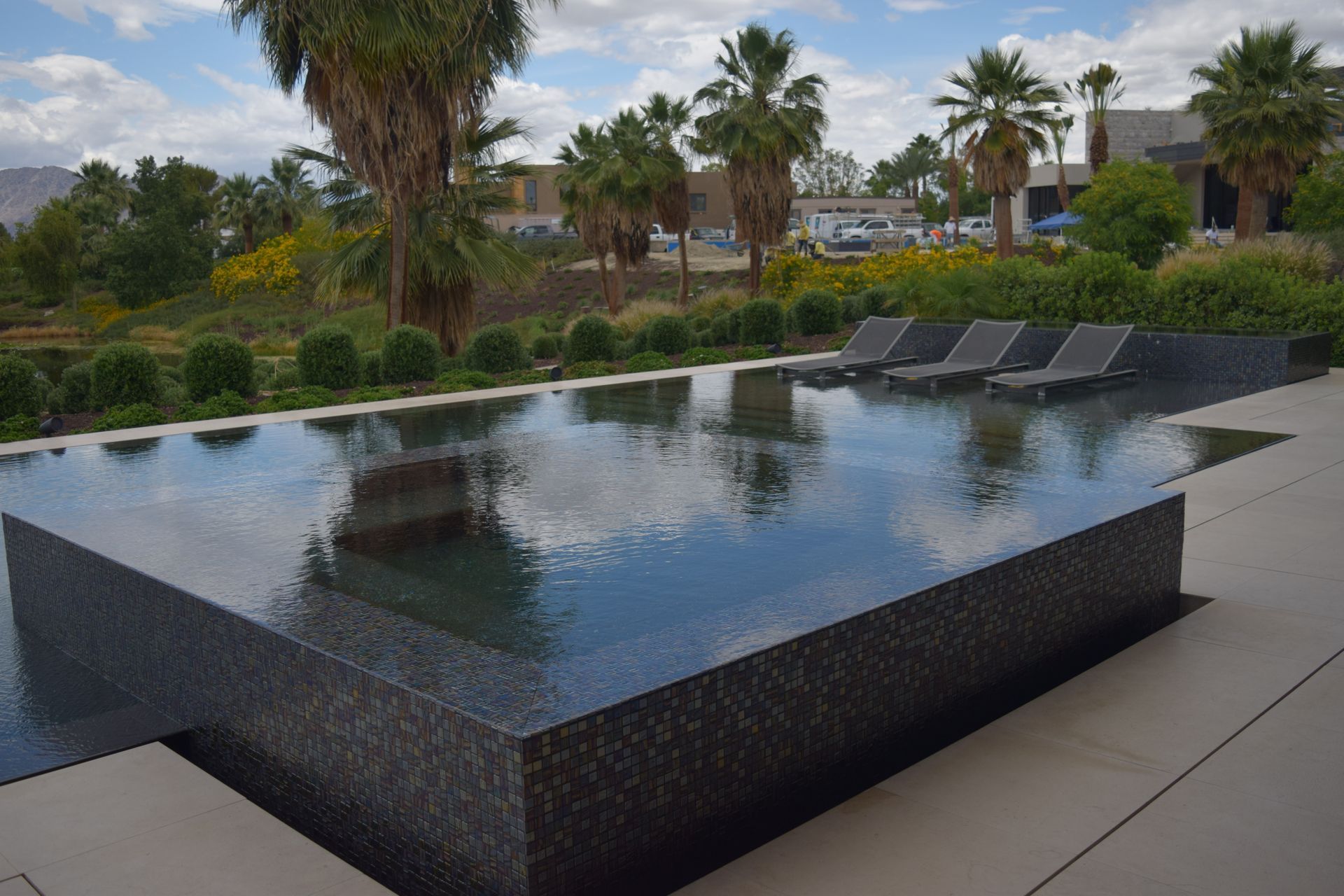 Rectangular pool with dark tile, reflecting sky. Lounge chairs, palm trees, and landscaping in background.