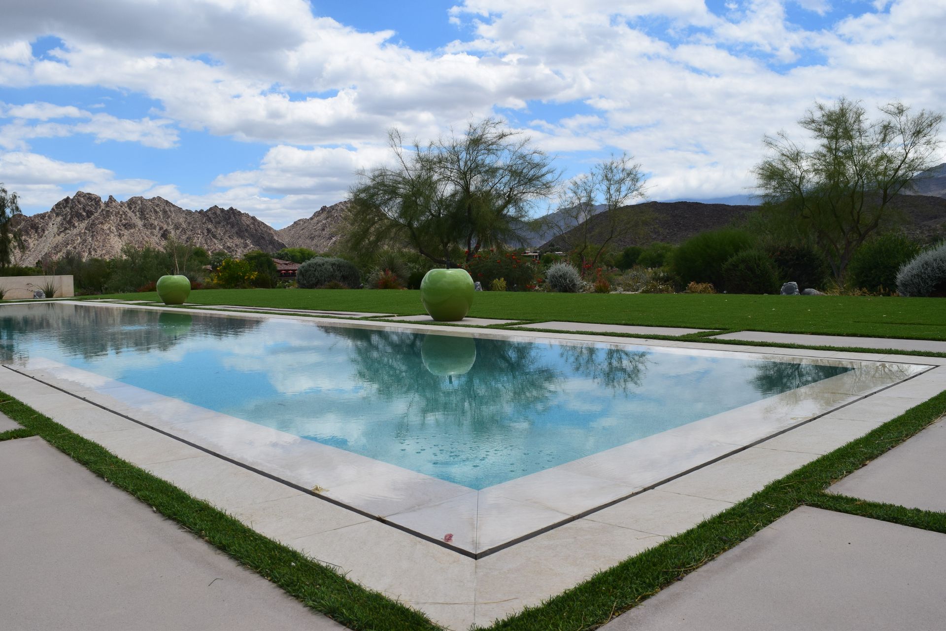 Modern backyard pool reflecting cloudy sky, with manicured shrubs and desert mountains in the background