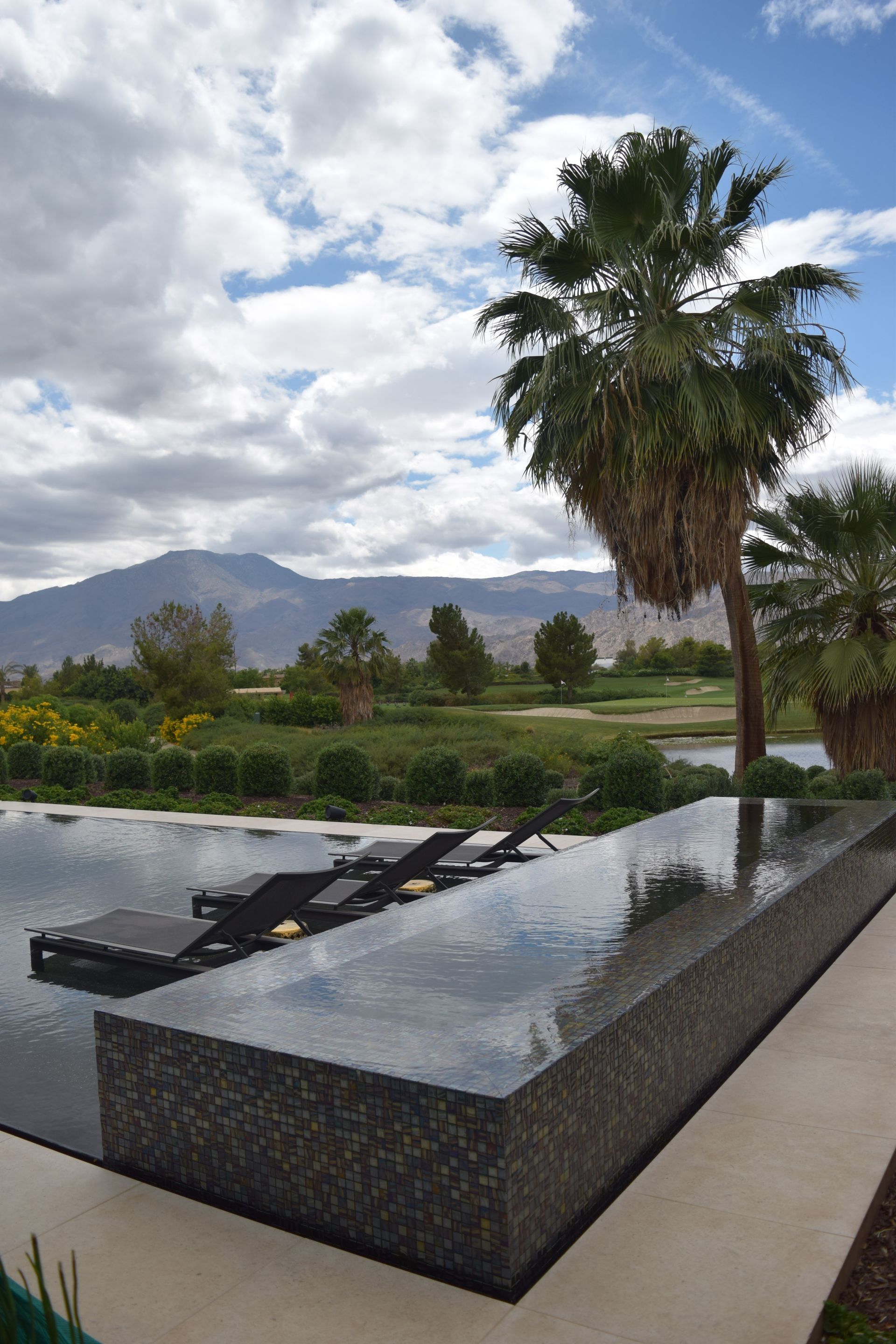 Infinity pool with lounge chairs, palm tree, and mountain view under cloudy sky.
