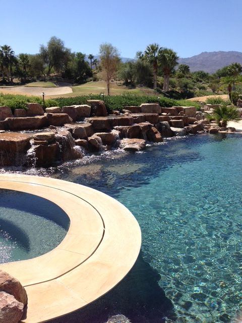 Curved pool beside a rocky waterfall, with clear blue water and desert hills in the background
