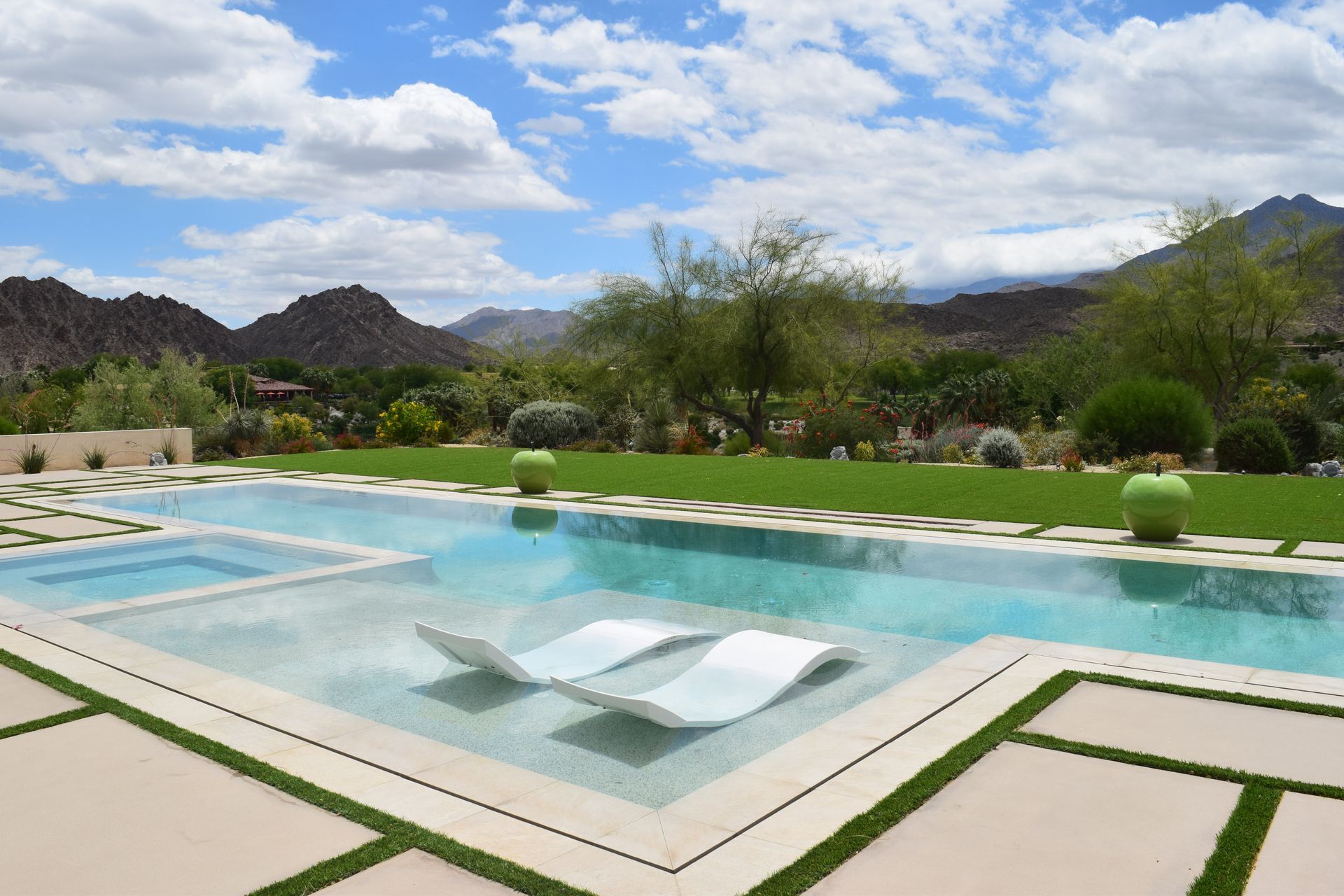 Swimming pool with floating chairs, lush lawn, and mountain backdrop under a partly cloudy sky.