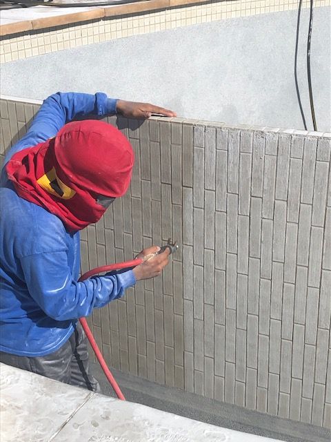 Worker in blue and red clothing tiles a gray wall with a trowel, using a red hose nearby.