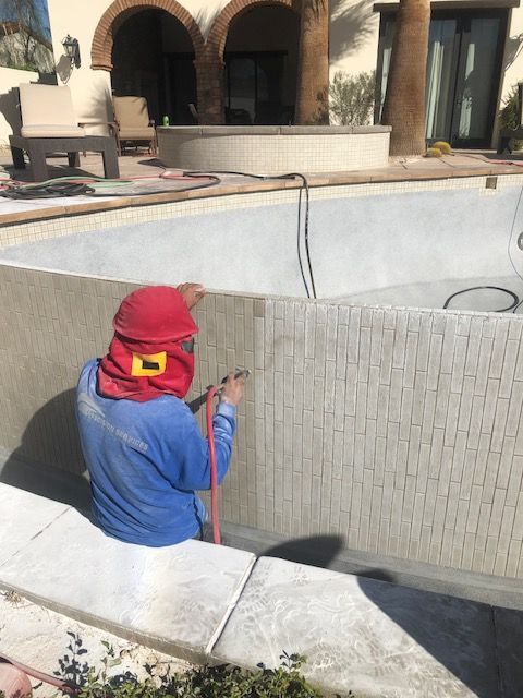 Worker in blue shirt and red hat sprays a pool wall beside a patio.