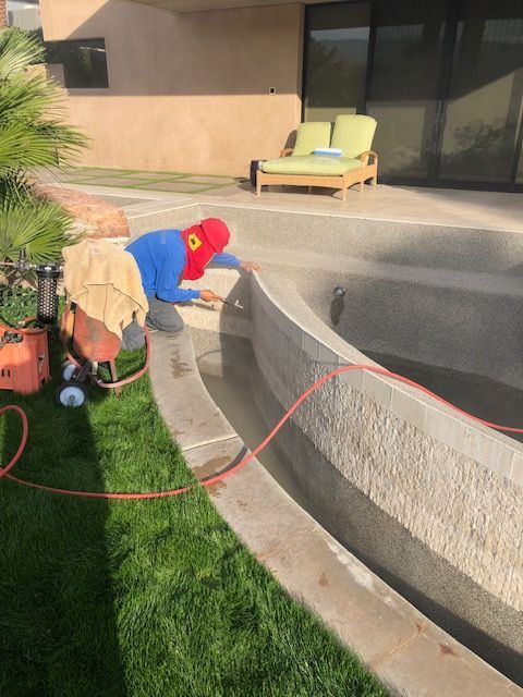 Person in red cap and blue shirt kneels by a backyard pool edge, washing the concrete with a hose.