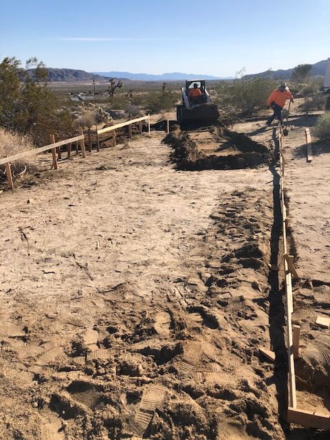 Construction crew grading a dirt path beside wooden forms in a sunny desert landscape