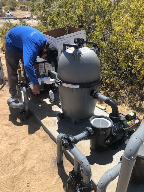 Worker in blue overalls bends over pool equipment beside a large gray filter tank outdoors.