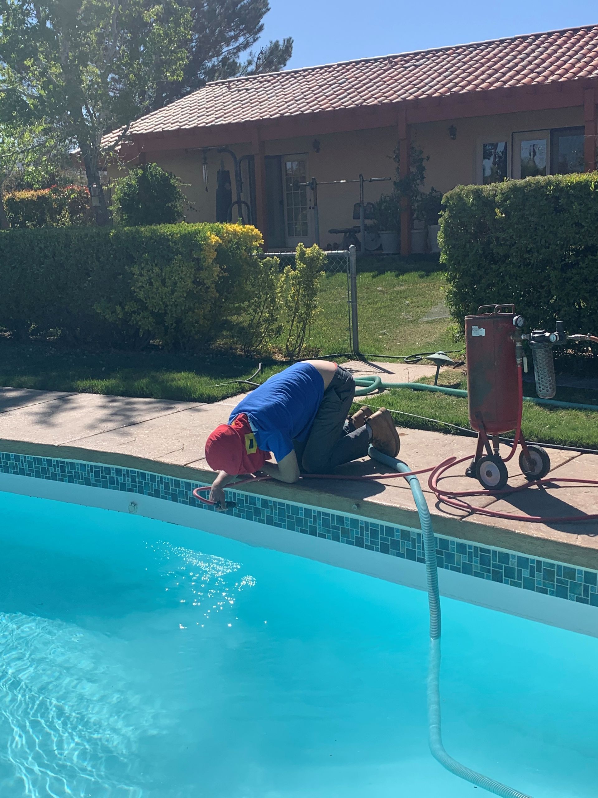 A person in a blue shirt and red cap kneels by a pool edge, using a red pressurized tank to clean the tile border.