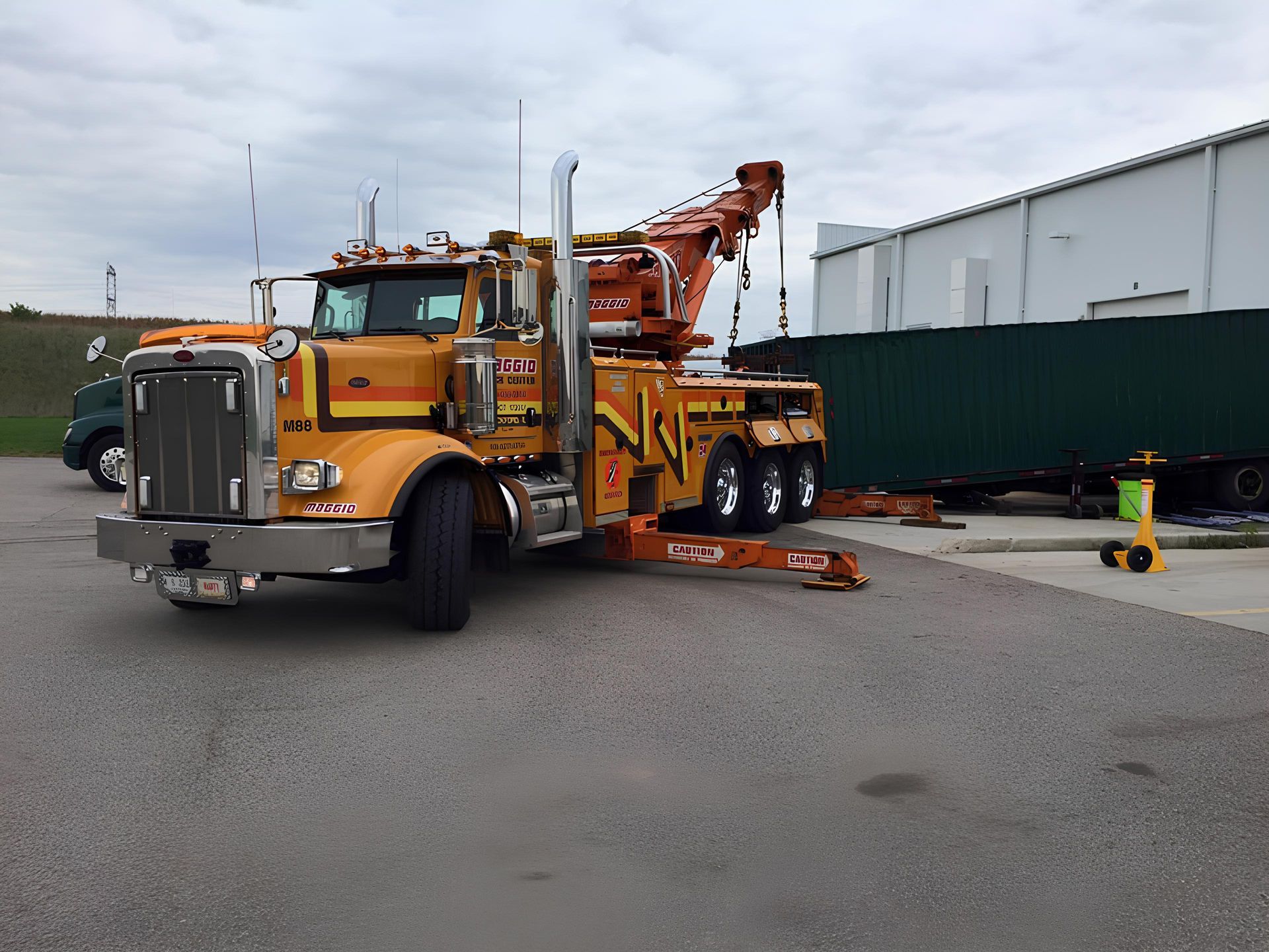 Yellow and orange heavy duty tow truck, parked with boom extended, preparing to lift a green container outside a white building.