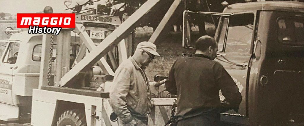 Two men near a tow truck. One is looking at something in his hand, the other at the truck. Black and white.