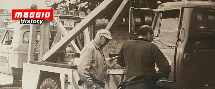 Two men near a tow truck. One is looking at something in his hand, the other at the truck. Black and white.