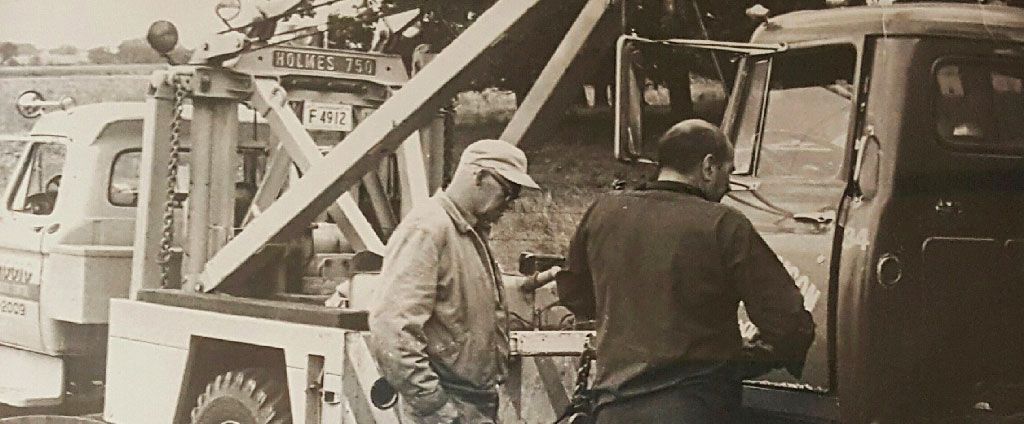 Two men near tow trucks, one looking at a device. Black and white photo.