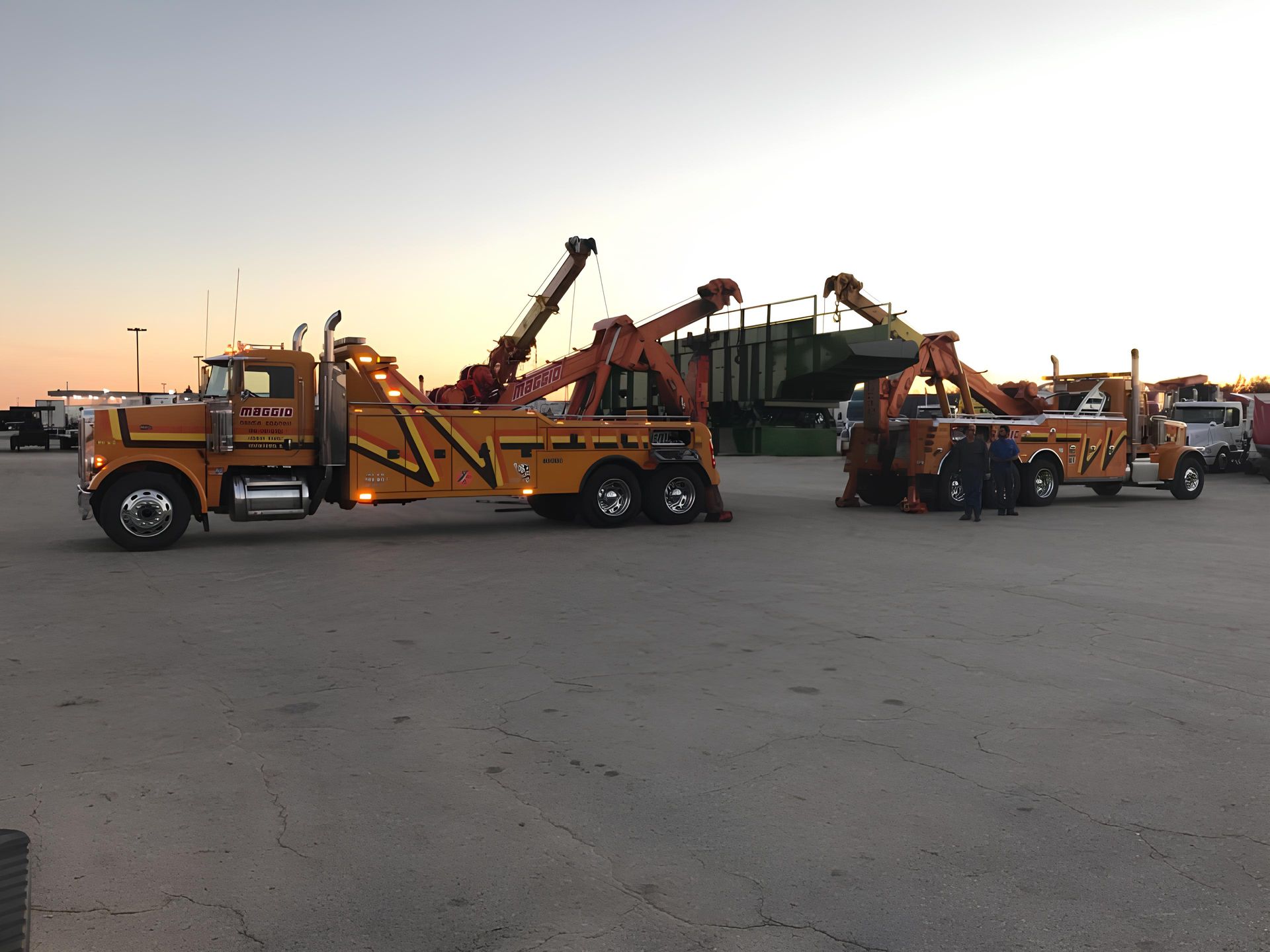 Two orange heavy duty tow trucks lifting a green object in a parking lot at dusk.