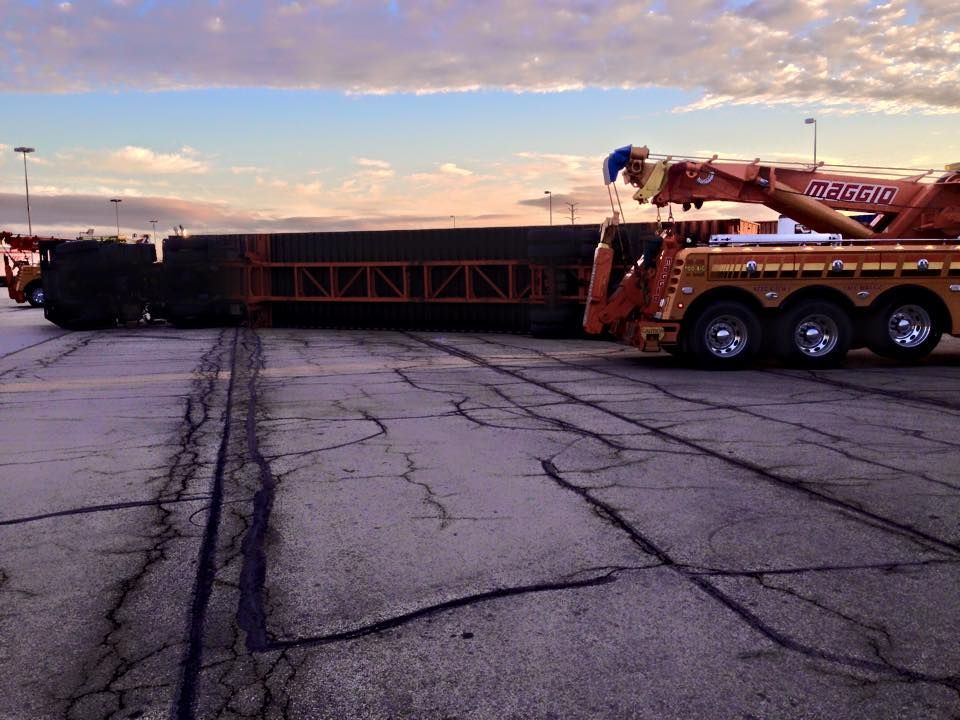 A tow truck lifting a tipped-over flatbed trailer in a cracked parking lot at dusk.
