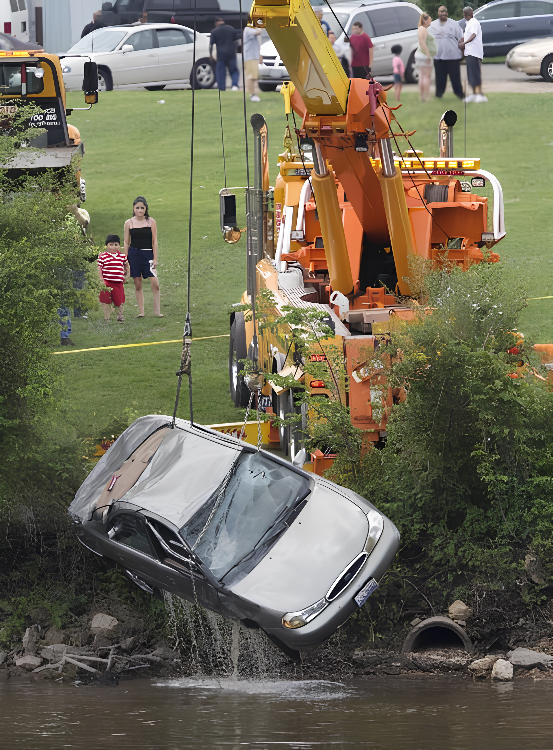 Tow truck lifting a silver car from a body of water; bystanders watch from a grassy area.