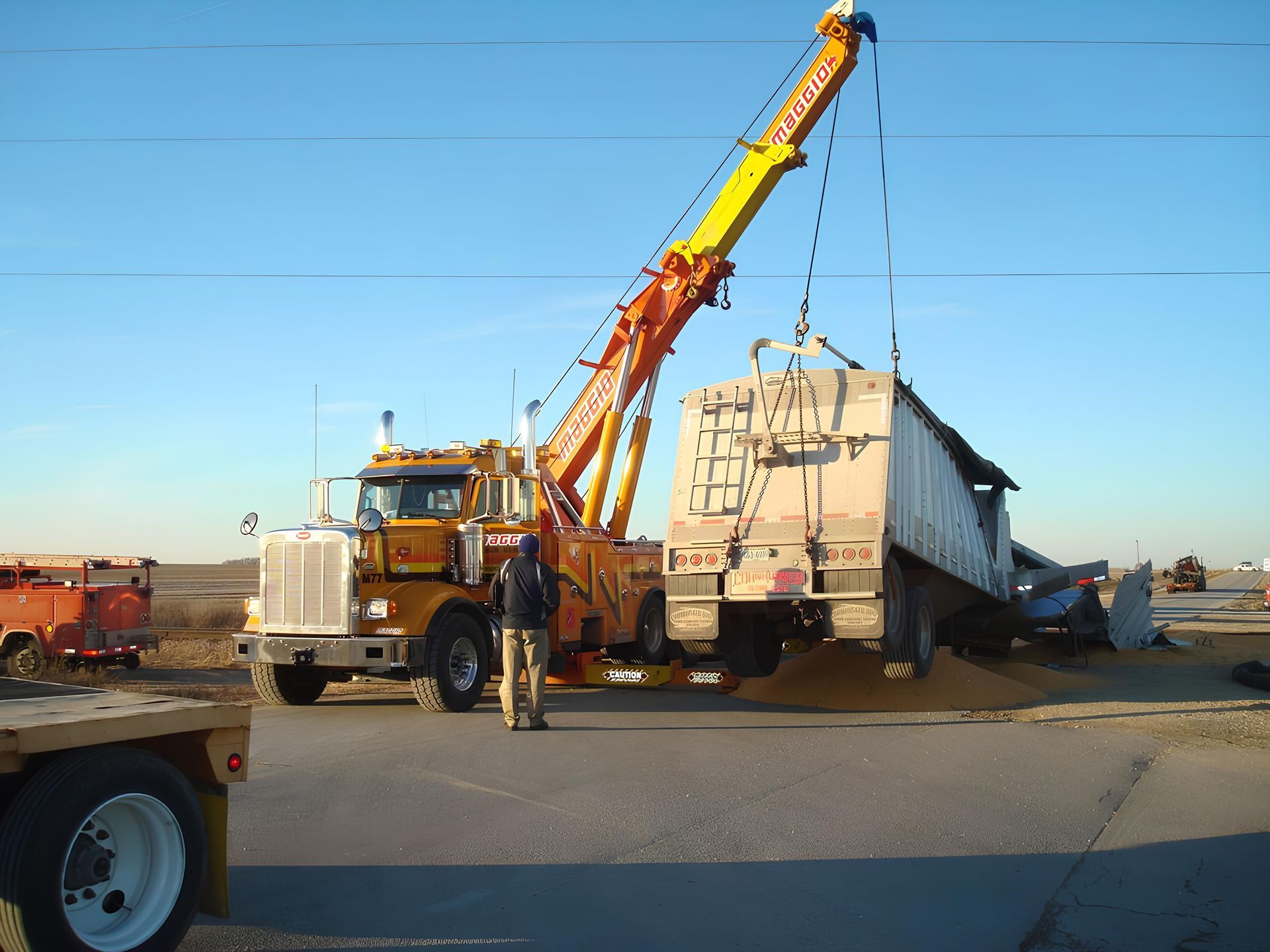 Tow truck lifting a damaged grain trailer on a road. Man stands nearby. Sunny day.