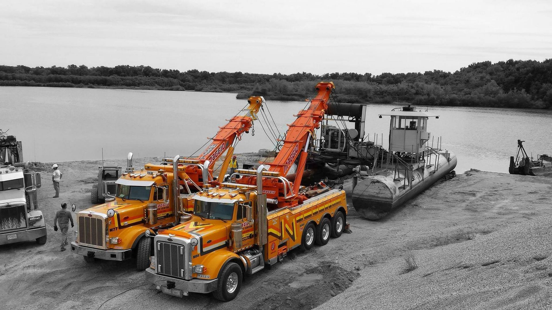 Two orange tow trucks near a barge on the shore. Monochrome background with trees and water.