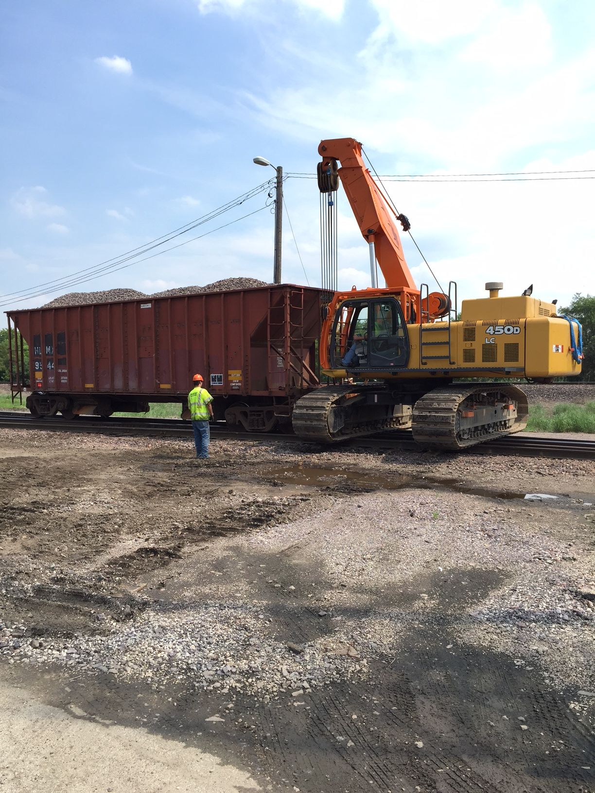 An excavator loading a rail car with material. A worker in a vest stands nearby on a gravel lot.