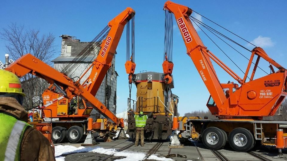 Two orange cranes lifting a yellow locomotive on a snowy train track; a person in a green vest stands between them.