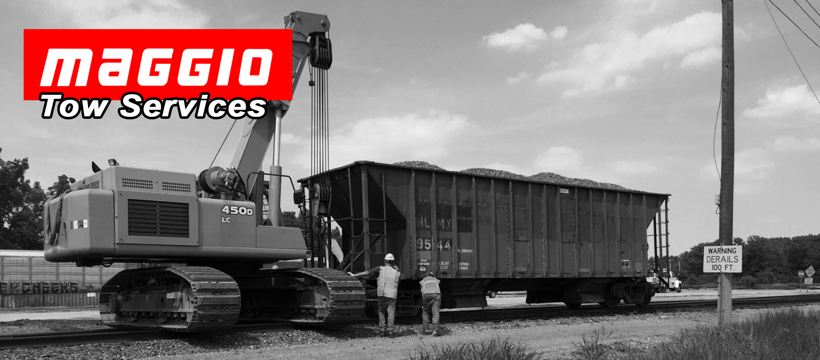 Excavator towing a rail car filled with cargo next to railroad tracks. Two people are near the car.
