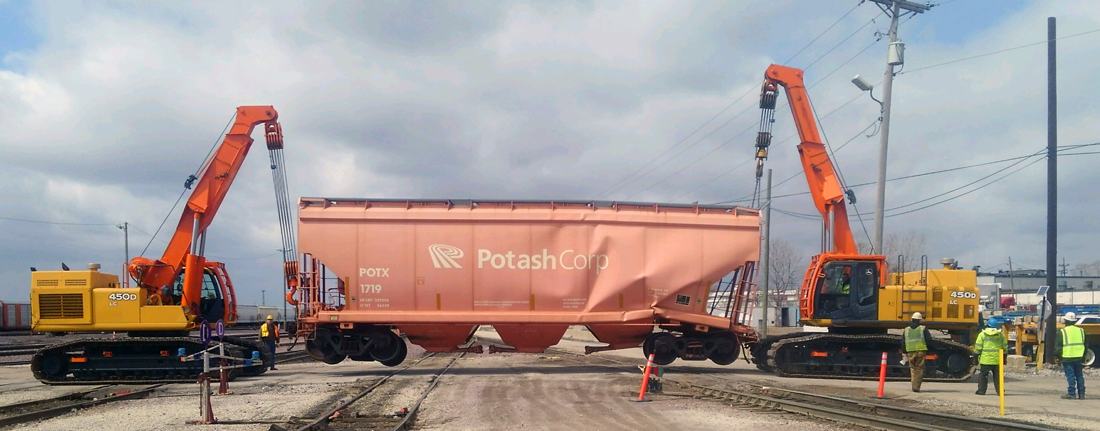 Two excavators lifting a damaged orange freight car on a railroad track under a cloudy sky.