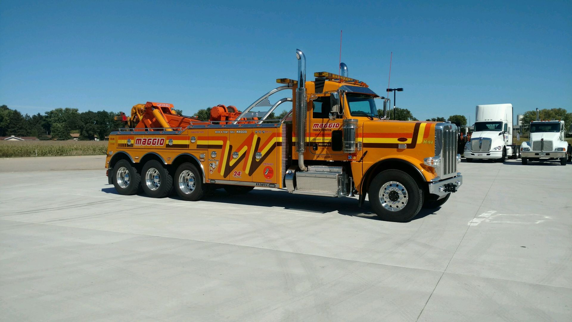 Yellow and orange tow truck on a concrete lot, with other trucks in the background under a clear sky.