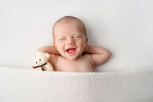Smiling baby lying in a white blanket with arms behind head, holding a small stuffed toy.