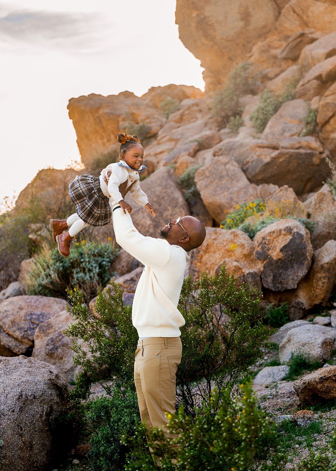 Man lifting a child in a white dress beside rocky desert cliffs and green shrubs.