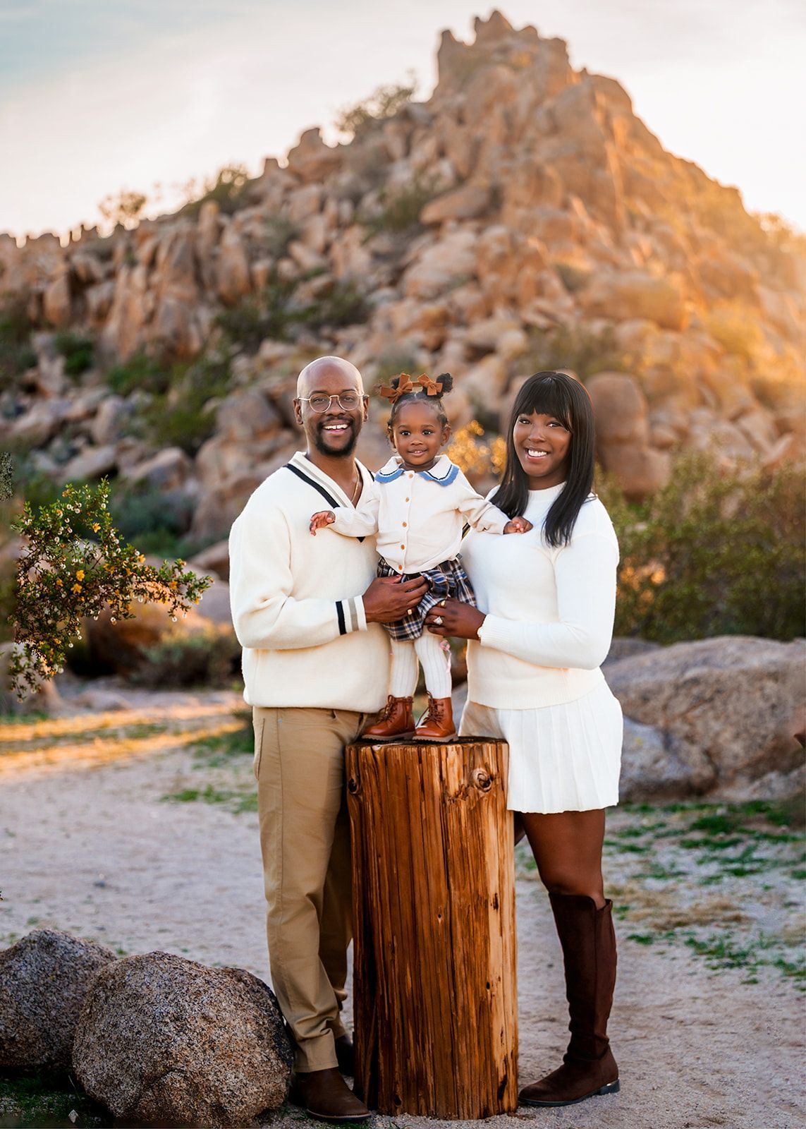 Family portrait outdoors by a rocky hillside, with a couple holding a smiling child on a tree-stump stand.