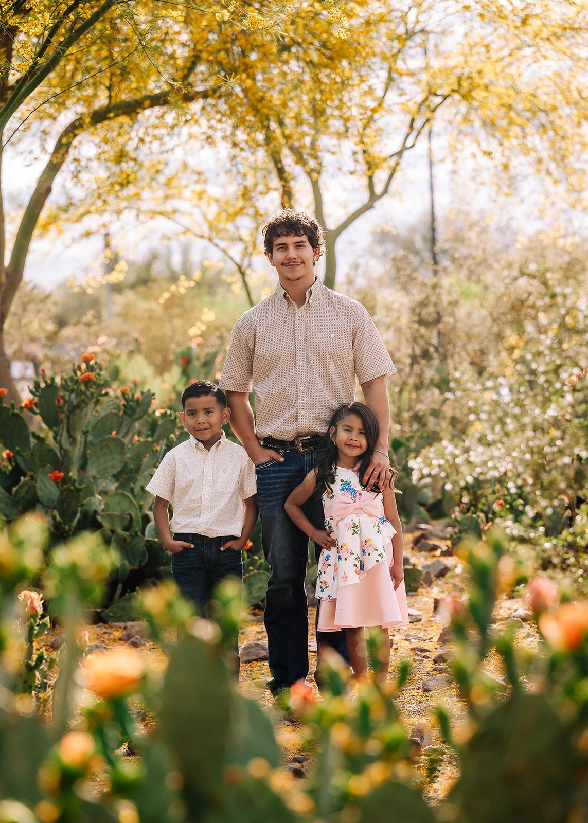 Three children standing in a flower-filled garden under yellow autumn trees.