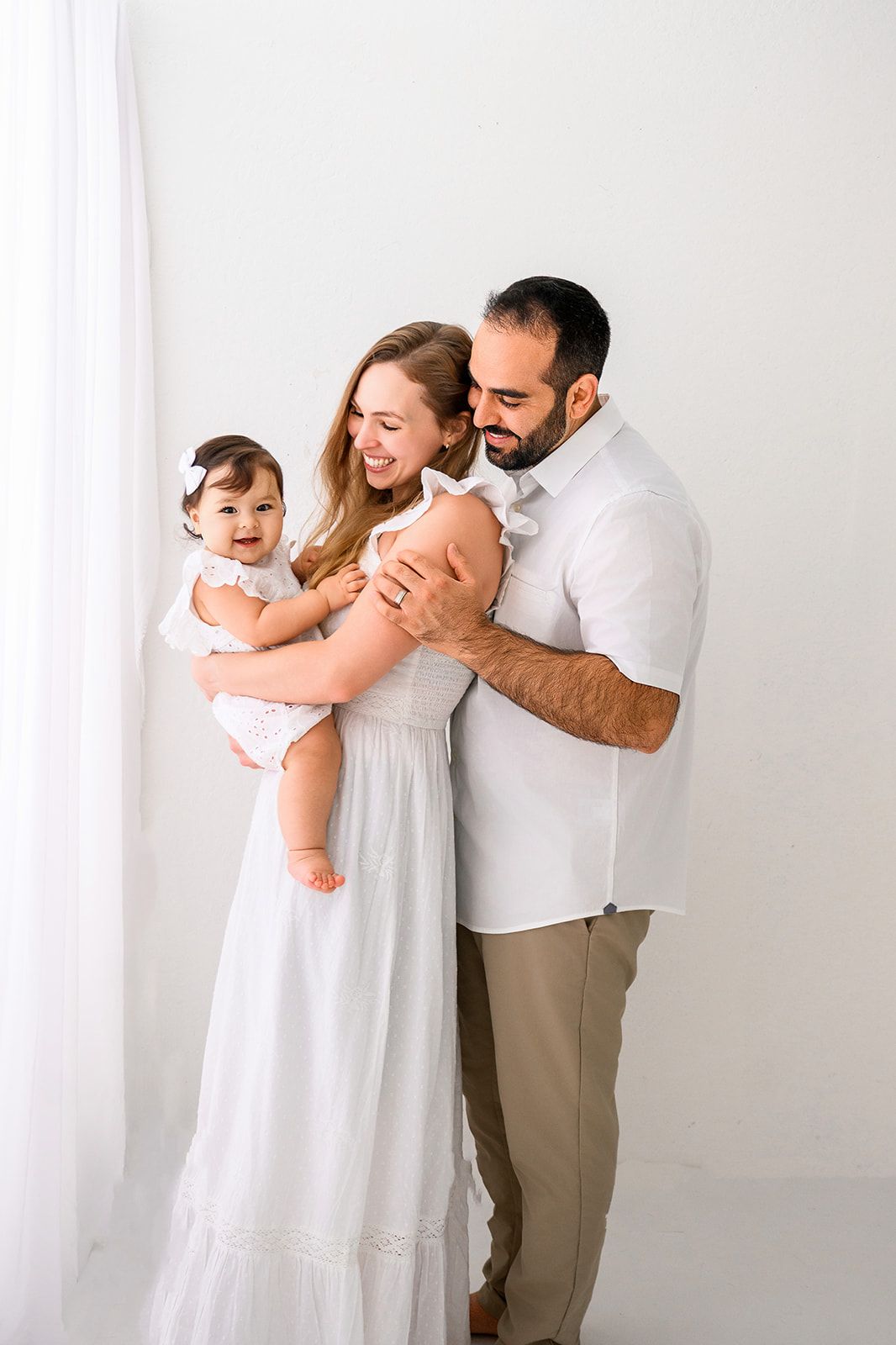 Family of three embracing in white clothing against a plain white backdrop.