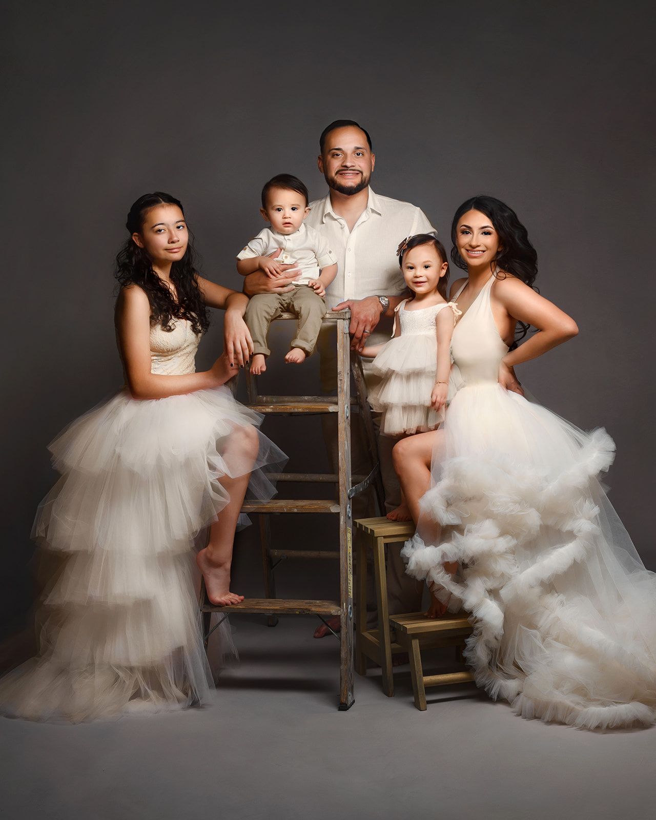 Family portrait with four children on stools, dressed in white tulle outfits against a dark studio backdrop.
