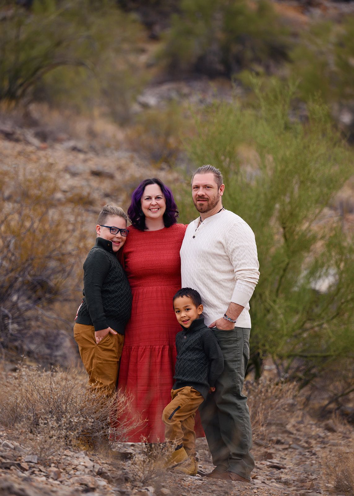 Family of four standing outdoors on a rocky hillside, with a woman in a red dress centered in the group.