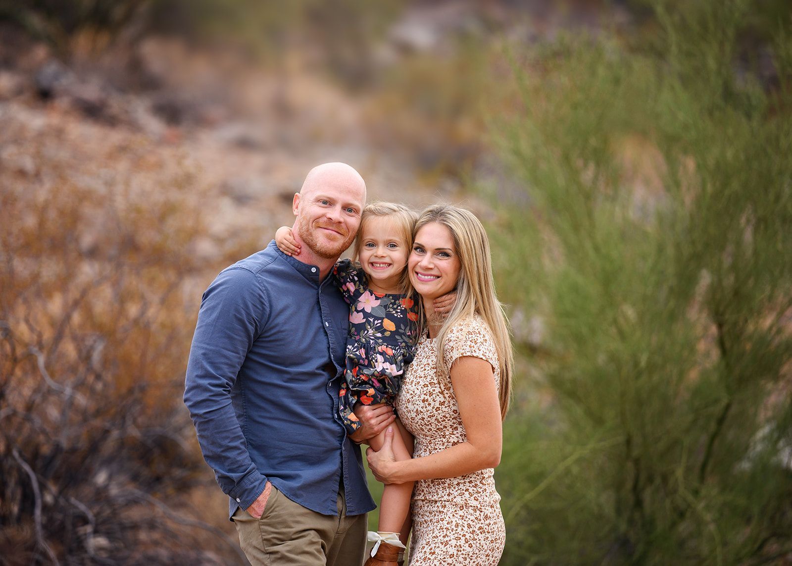 Three people smiling outdoors in a natural setting, with dry brush and a blurred background.