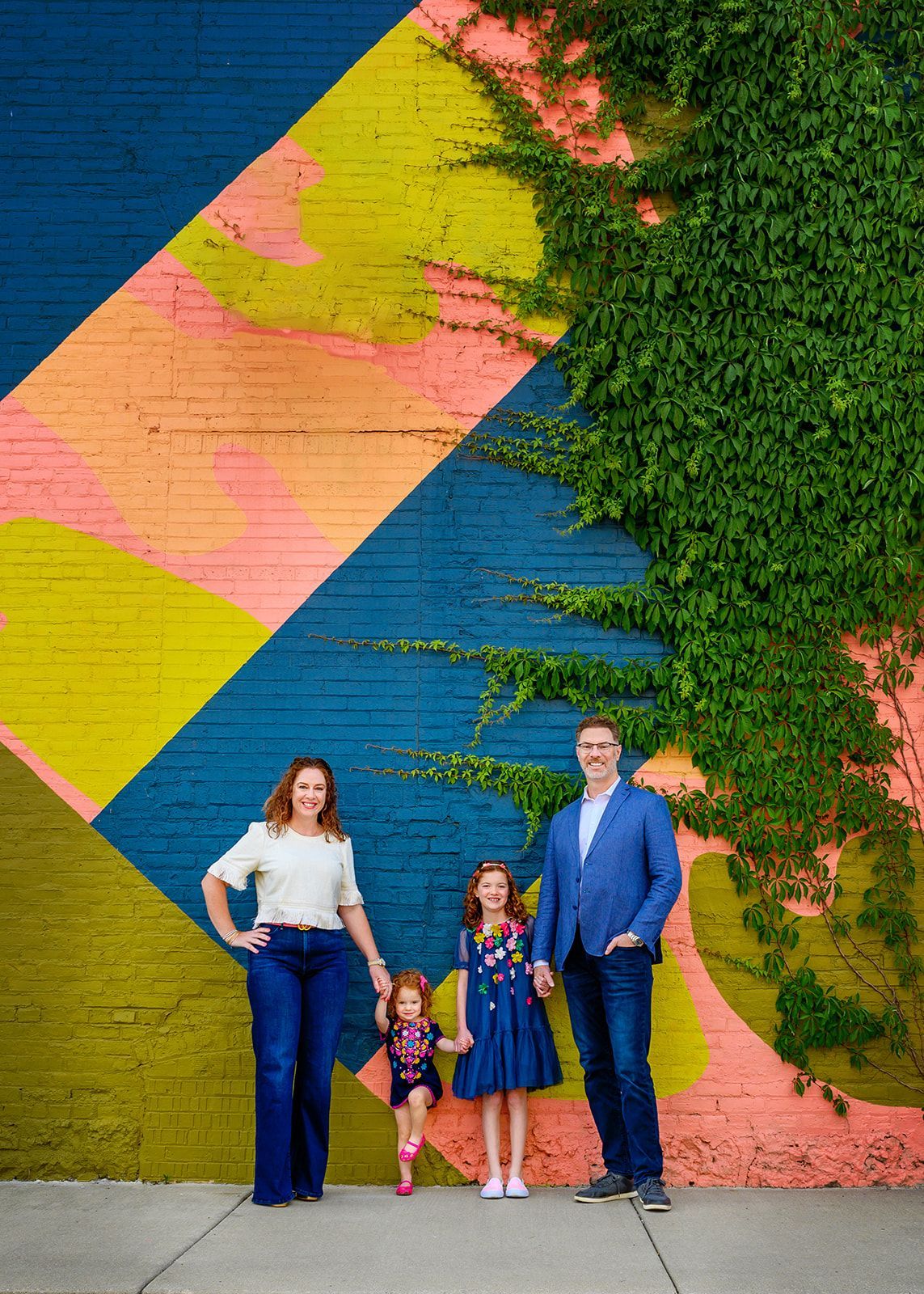 Family of four posing in front of a colorful geometric mural and greenery.