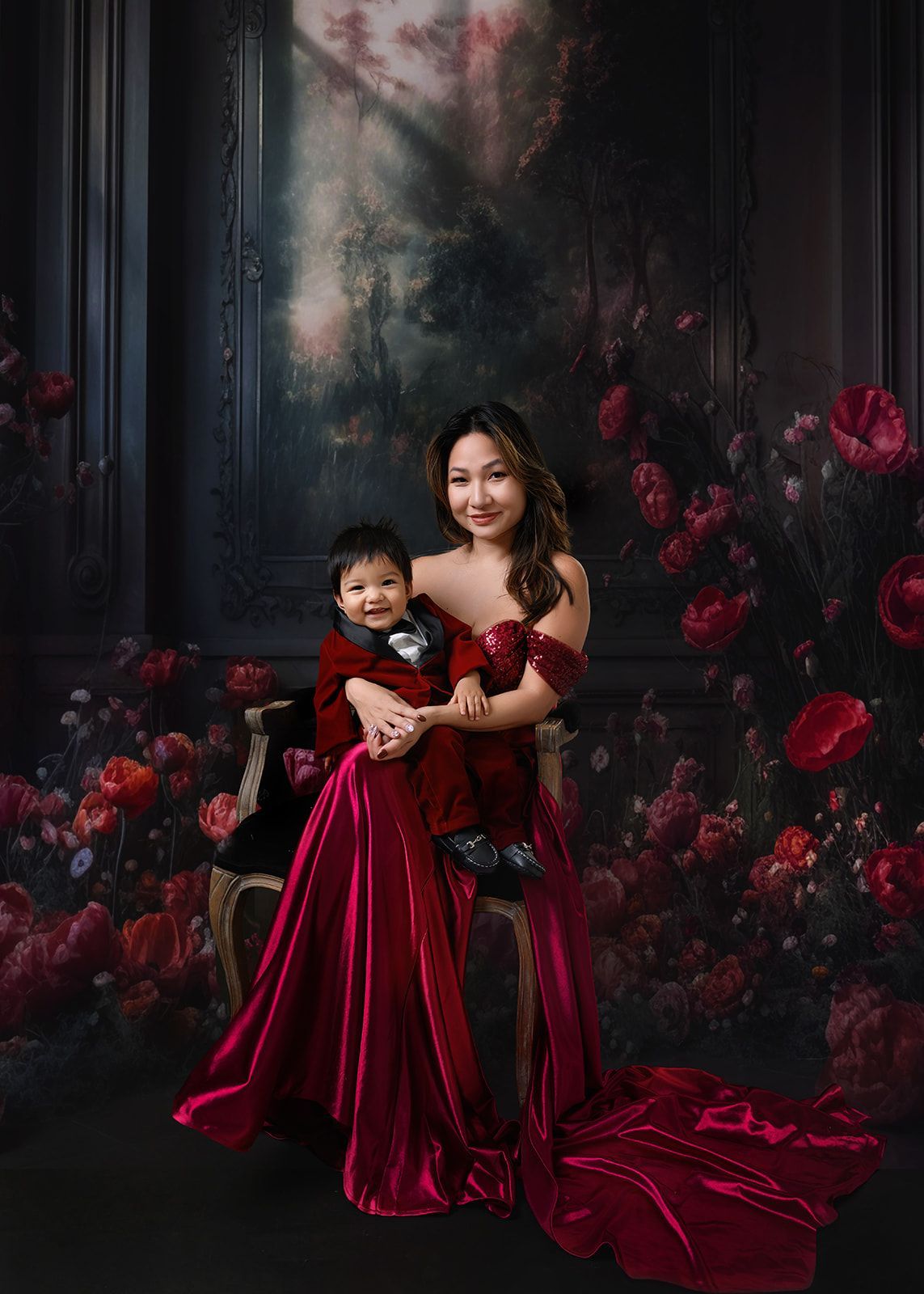 Woman holding a child in matching red dresses against a dark floral backdrop.