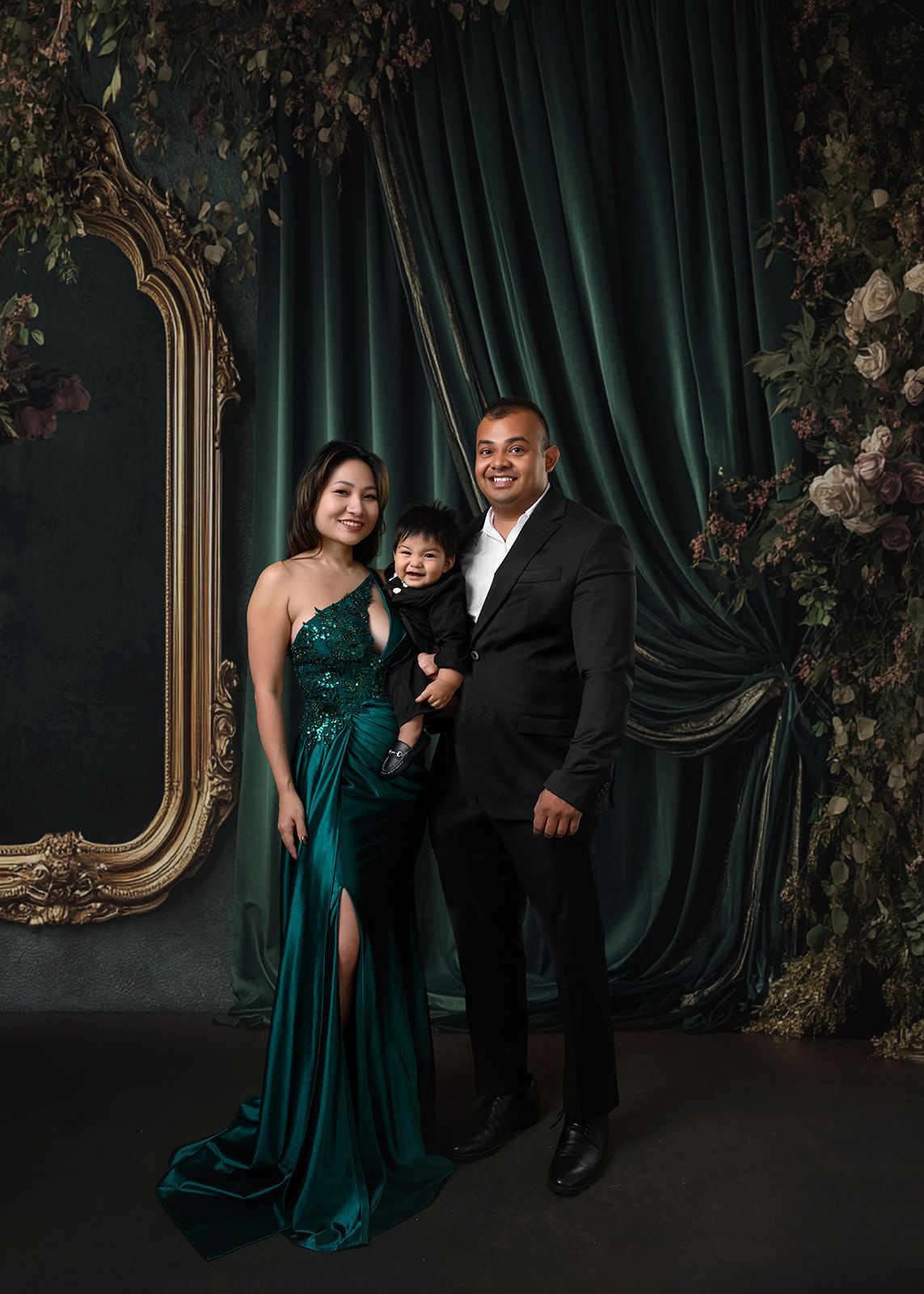 Three people posing in elegant formalwear before dark velvet drapes and an ornate floral backdrop.