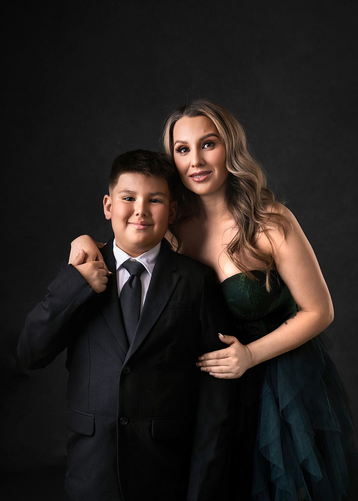 Two people posing in formal black attire against a dark studio backdrop, smiling gently.