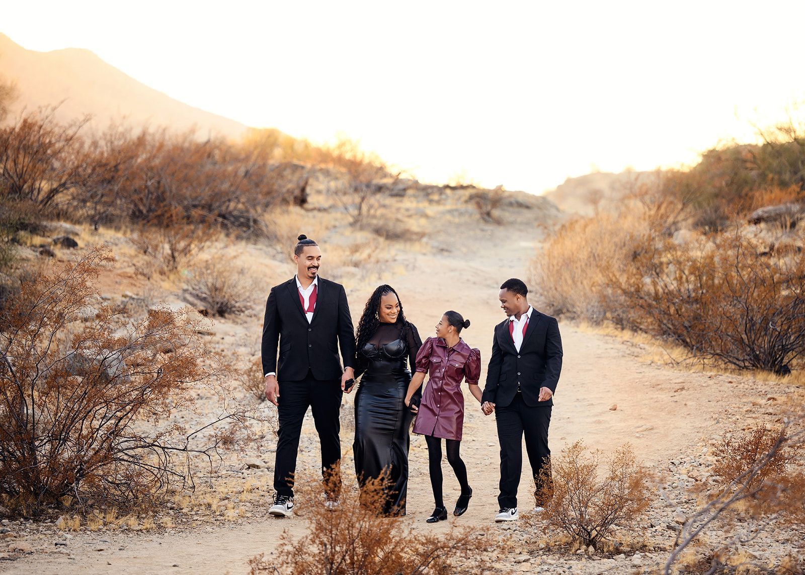 Four people in dark clothing walking together on a sandy desert trail at sunset.