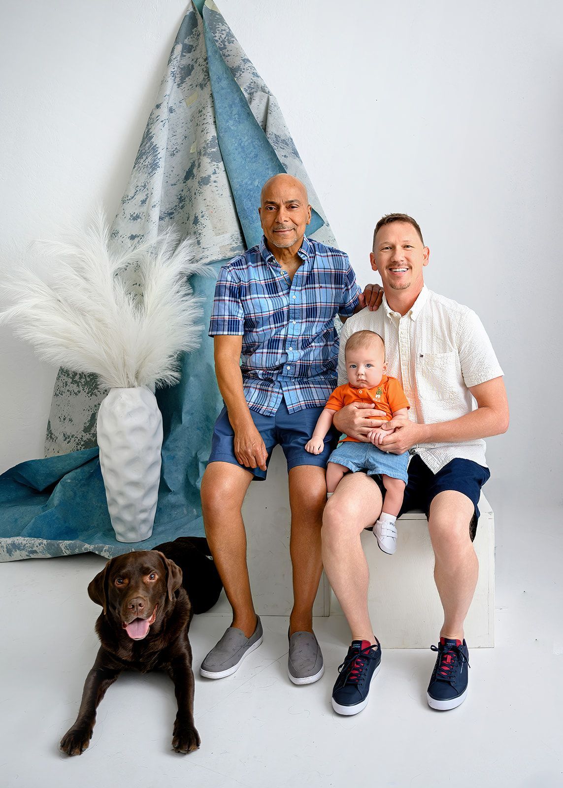 Two men and a baby seated with a dog in a studio, beside blue decor and a white vase.