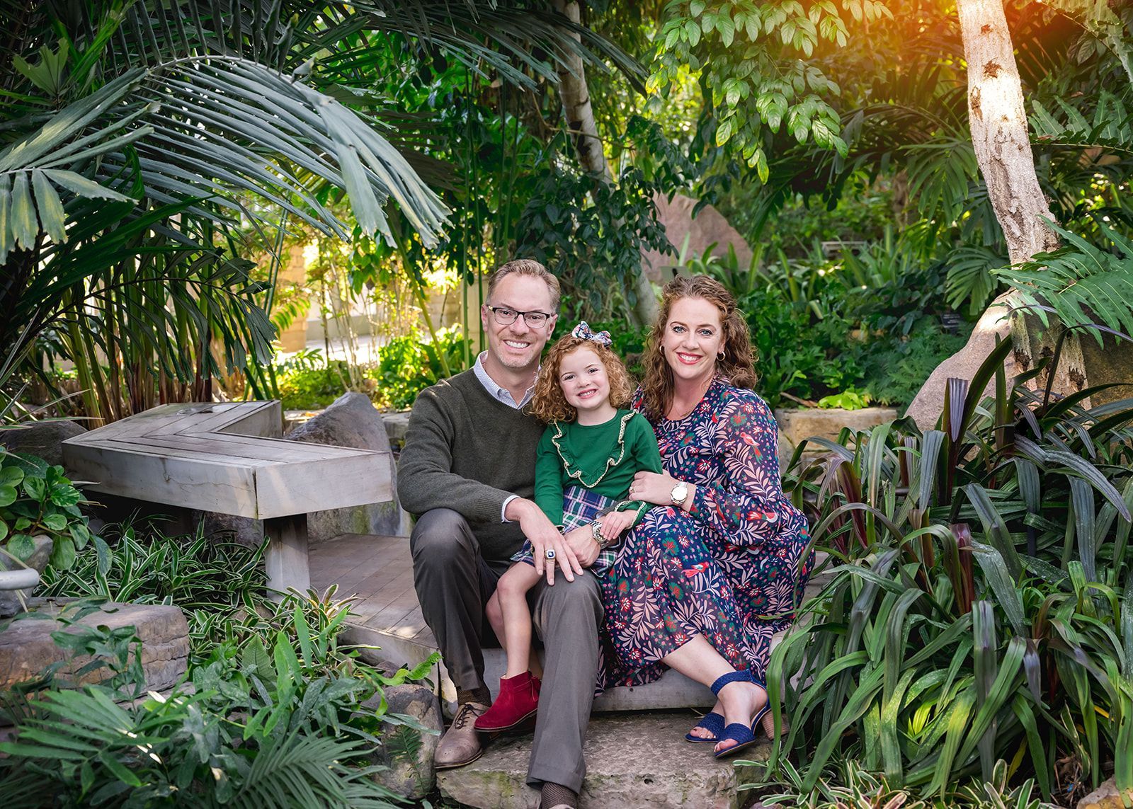 Family seated on a bench in a lush tropical garden, smiling at the camera.