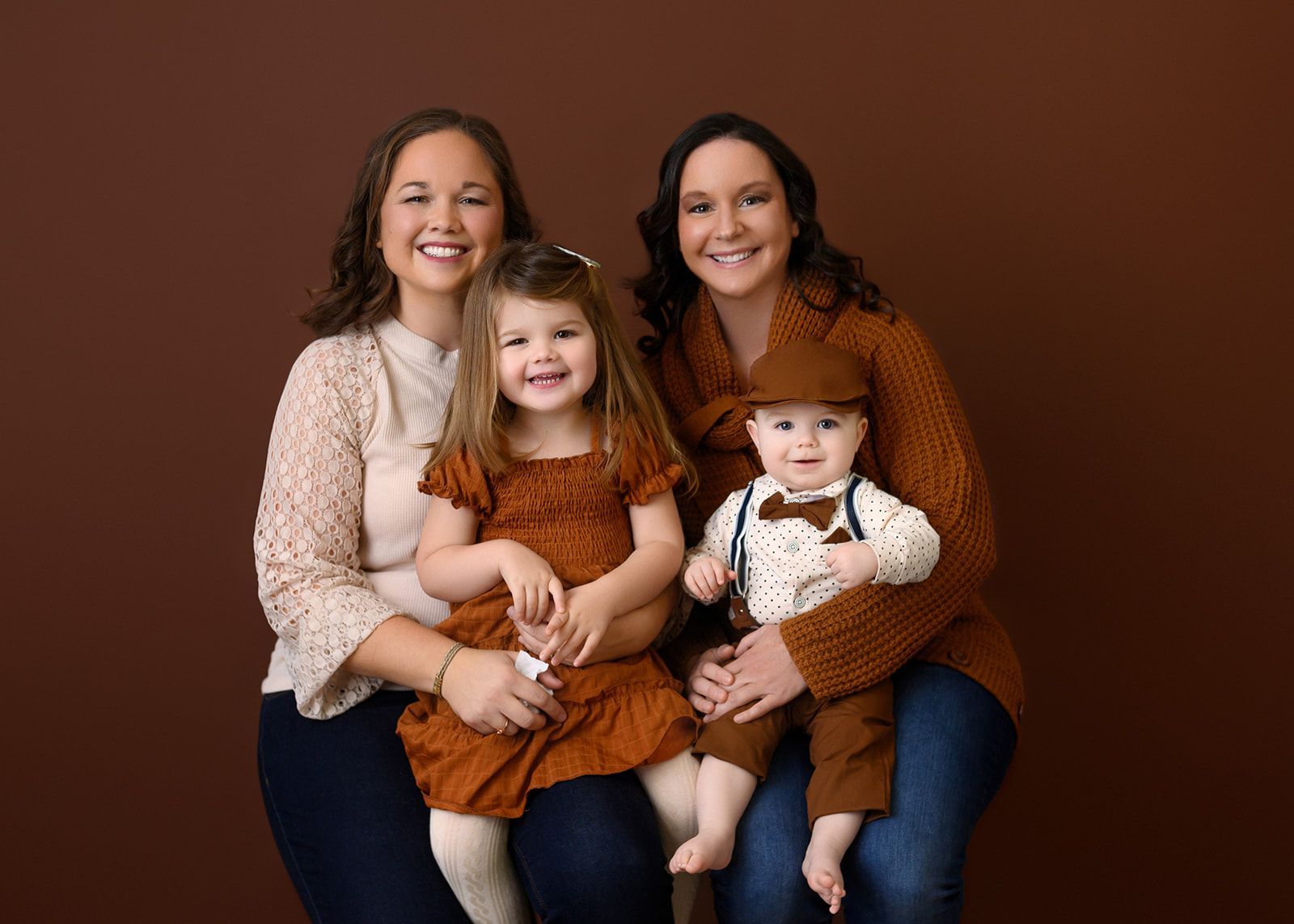 Four smiling people seated together against a brown studio backdrop, wearing matching rust and cream outfits.