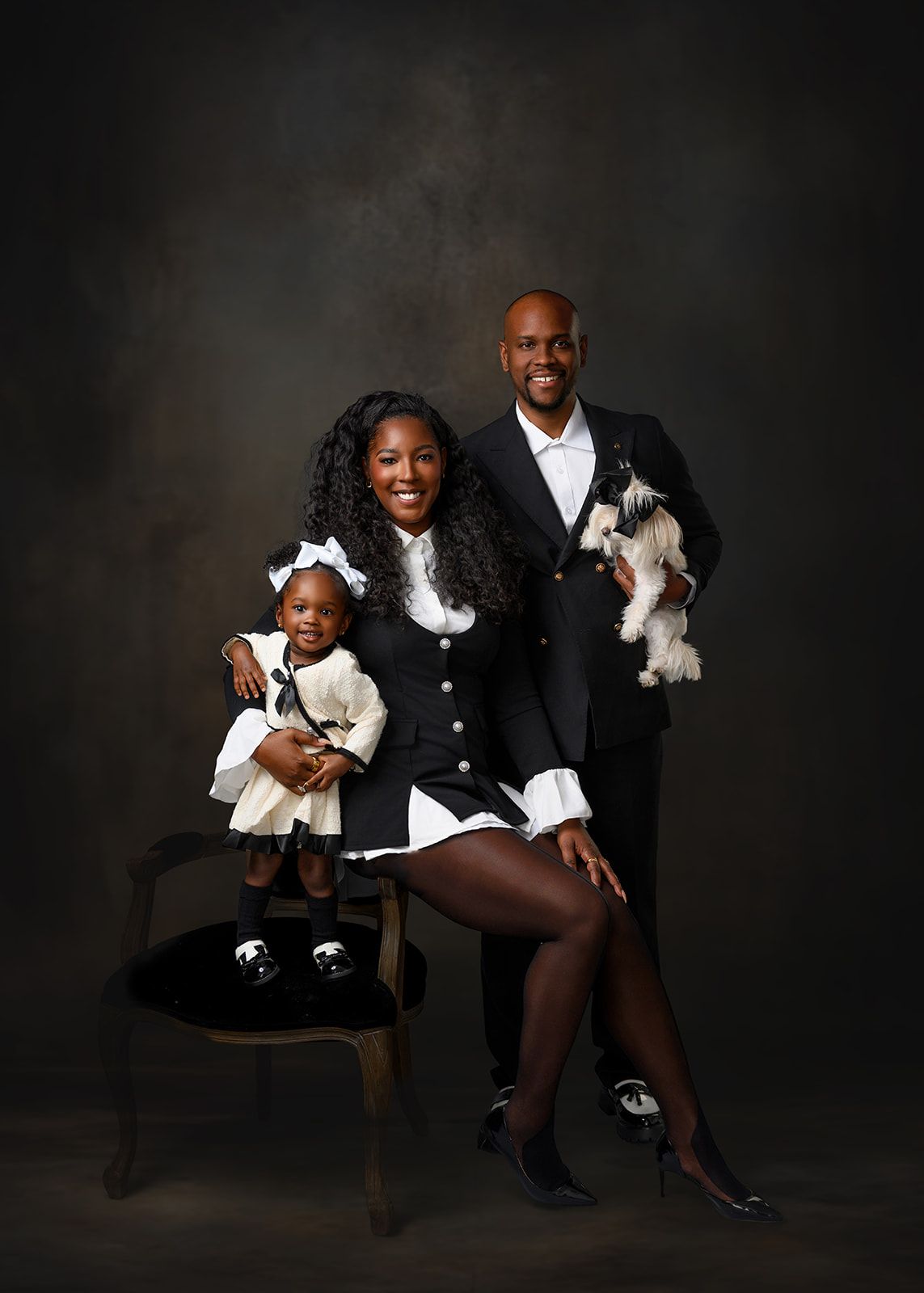 Formal family portrait of three people and two small dogs in black-and-white outfits on a dark studio backdrop.