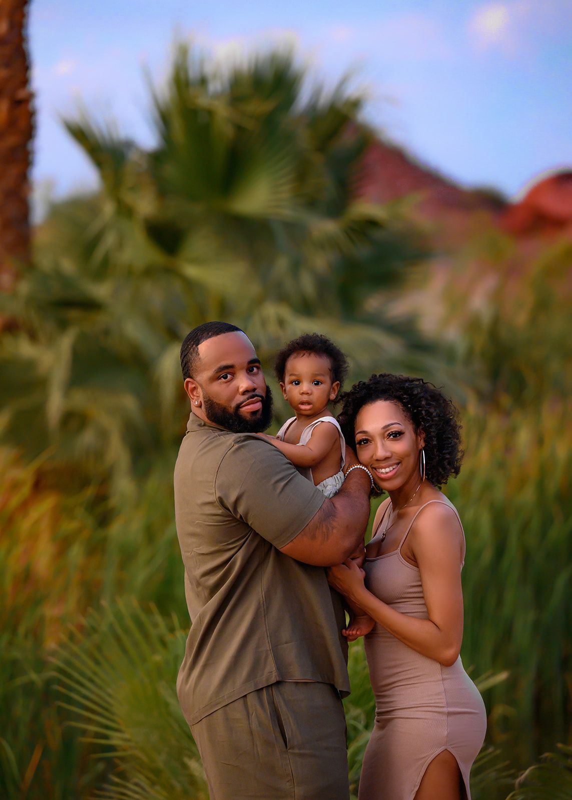 Family posing outdoors in a field, with blurred palm trees and warm sunset light.
