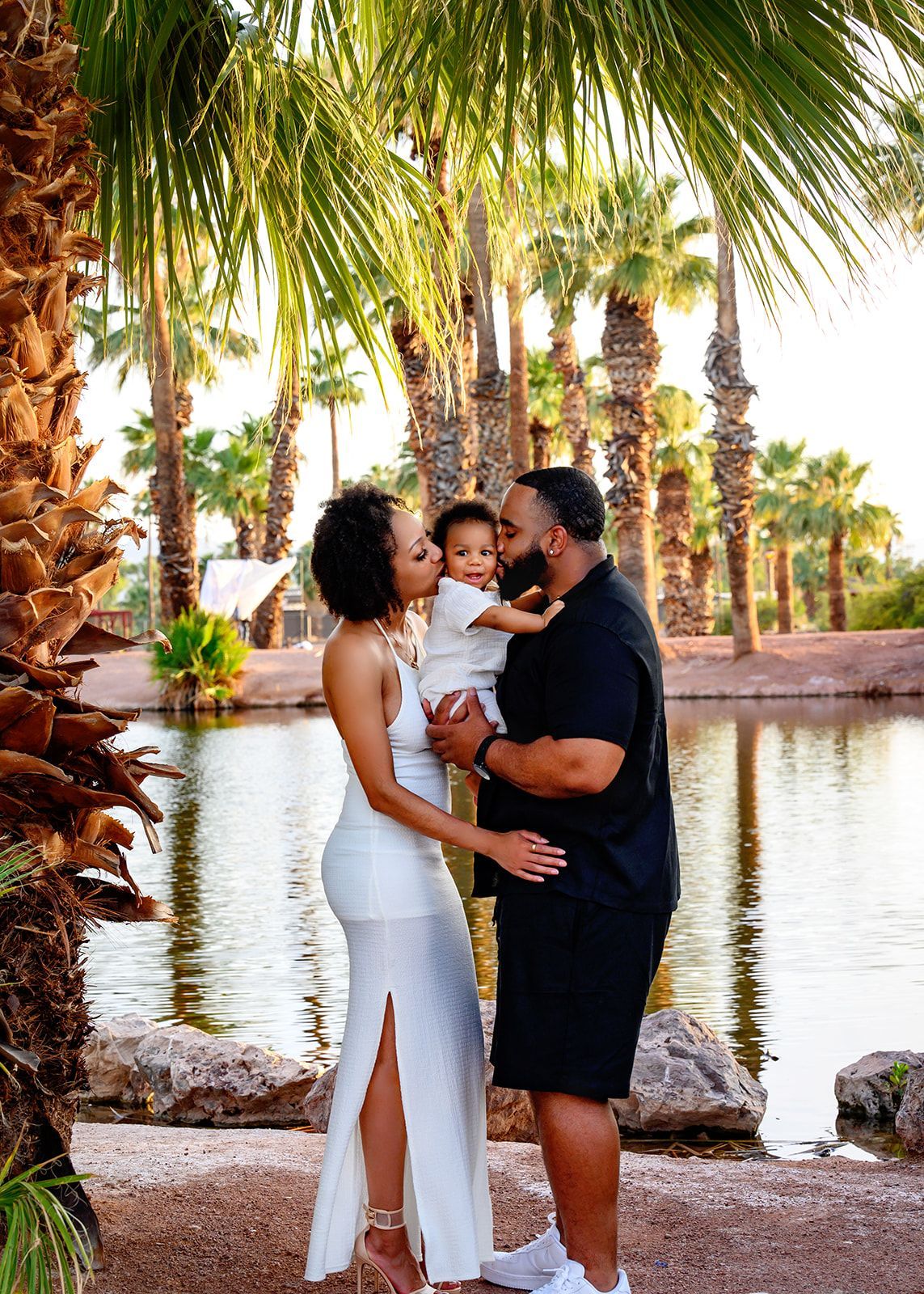 Family posing by a palm-lined pool, with a man holding a child beside a woman in white.