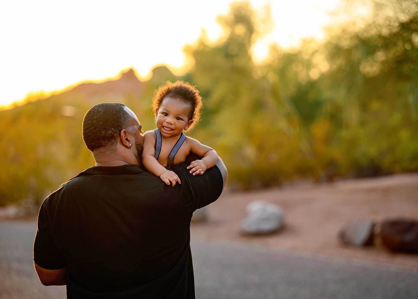Adult holding smiling child outdoors at sunset, with warm golden light and blurred trees in the background.