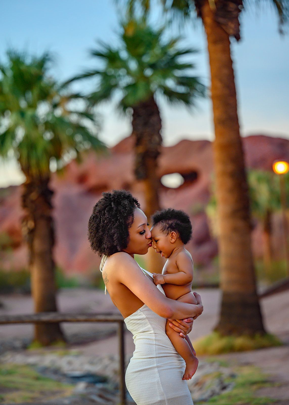 Woman holding a toddler outdoors at sunset with palm trees in the background.