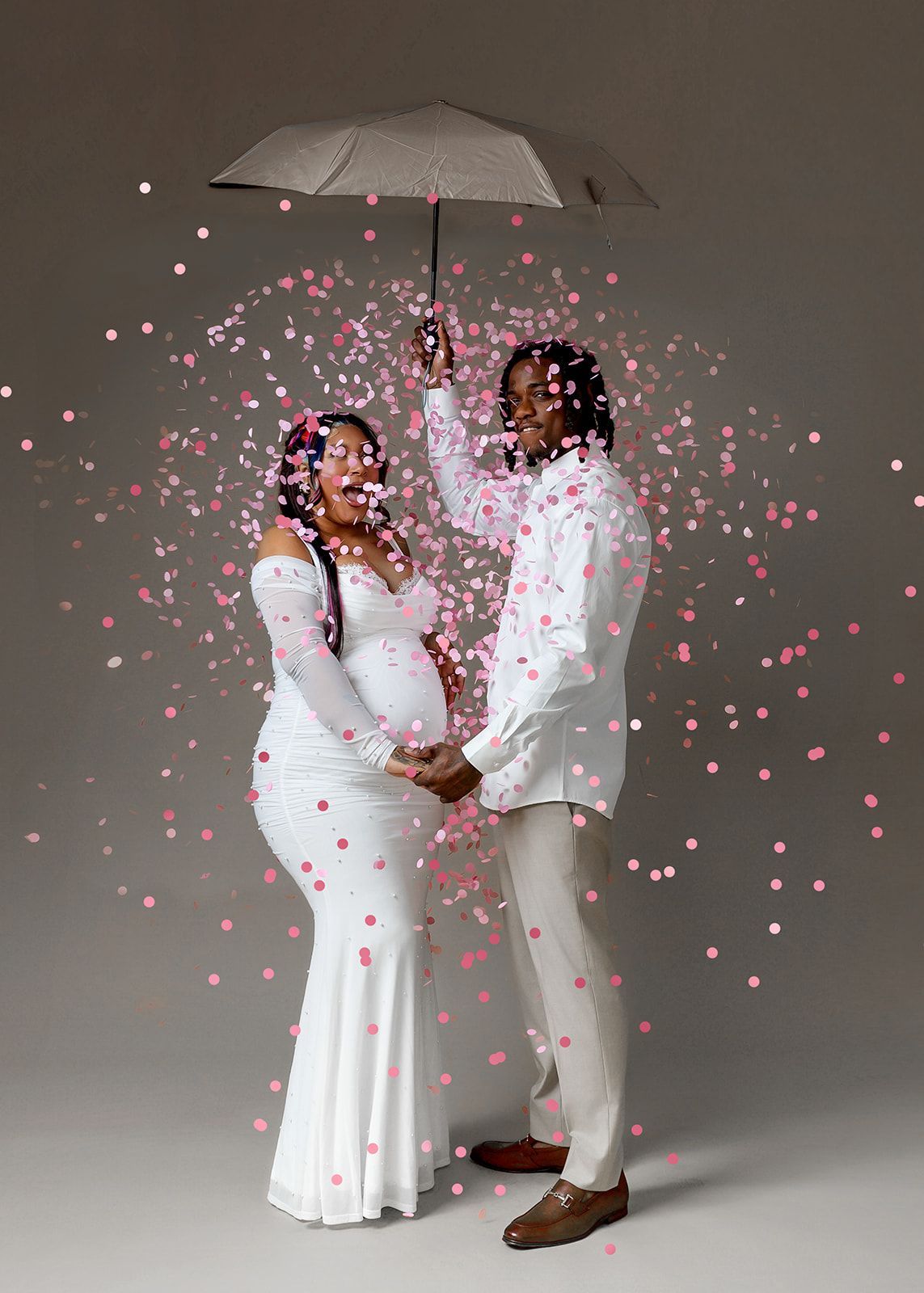 Couple under a white umbrella amid pink confetti, dressed in white against a gray backdrop.