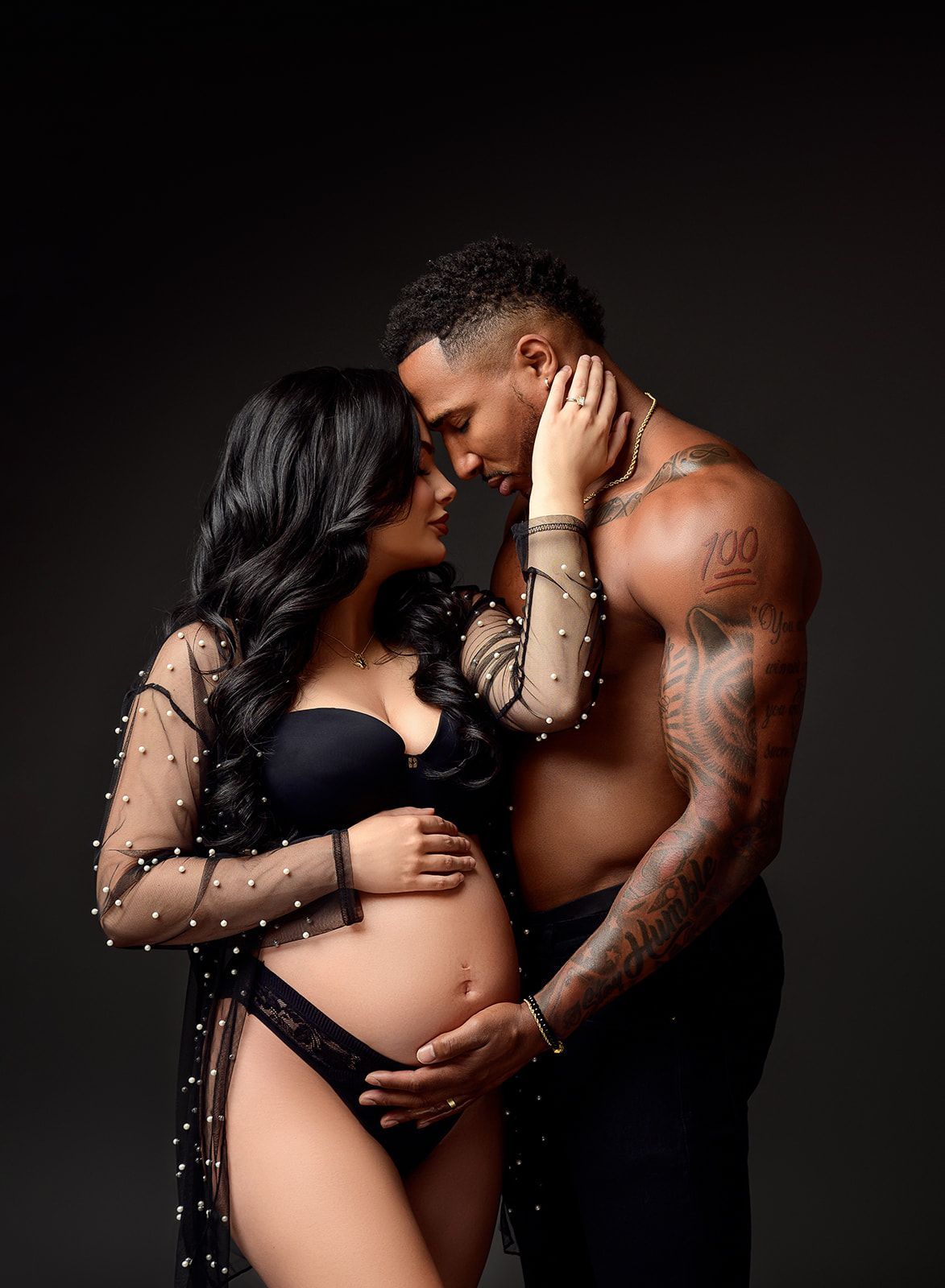 Couple in black lingerie and jeans embracing against a dark background, pregnant belly visible