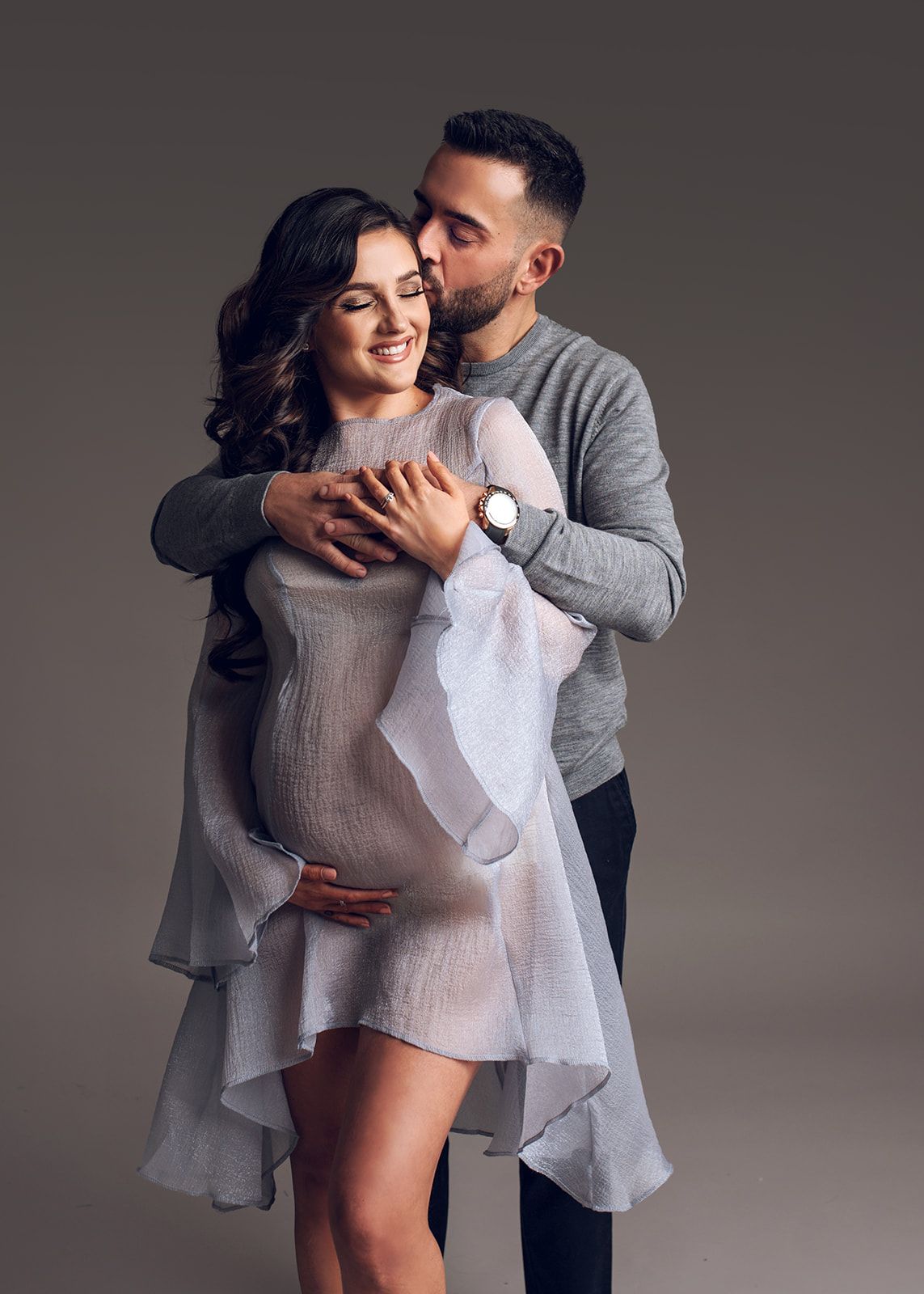 Couple embracing and smiling in gray outfits against a dark studio background